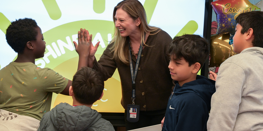An educator wearing a staff badge exchanges a high-five with a student in front of a display featuring the word “SHINE.” Several other students stand nearby, waiting their turn. A congratulatory balloon is visible in the background, and the indoor school setting is brightly lit.