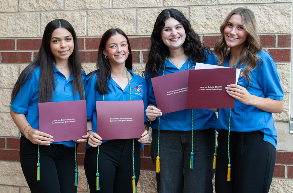 Four students wearing blue Teacher Cadet shirts stand in front of a brick wall, smiling and holding certificates.