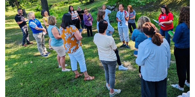 Group of adults standing in a circle outdoors on a sunny day, engaged in conversation in a green park area with trees.