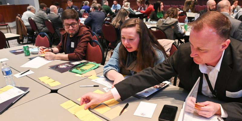 People seated at tables in a conference room, engaging in a group activity with sticky notes and papers spread out.