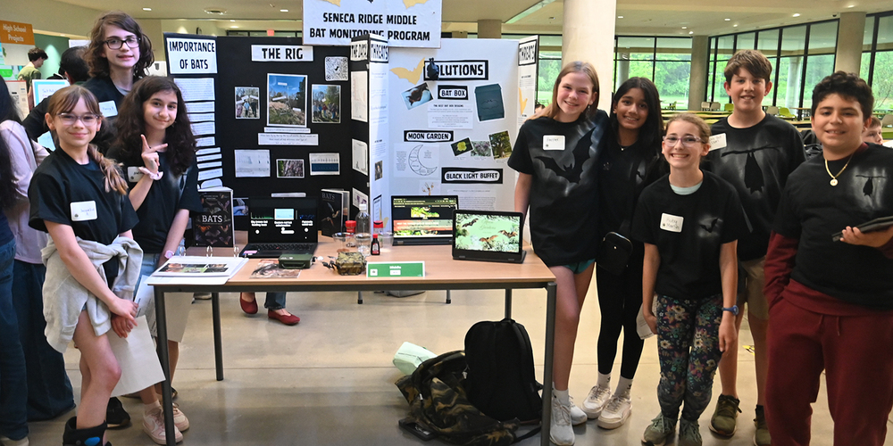 Group of students standing around a table displaying a science project about bat conservation, with posters, photos, and laptops, and a central sign reading “Seneca Ridge Middle Bat Monitoring Program.”