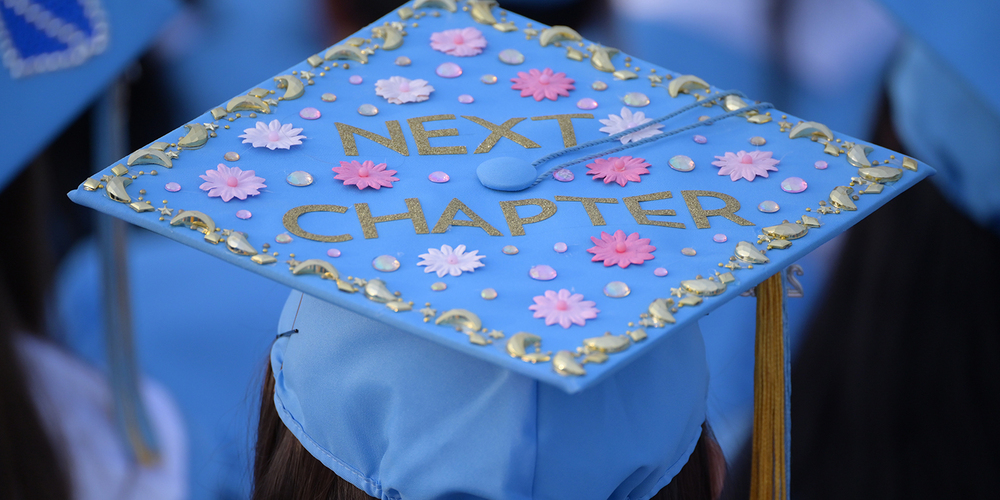 A close-up view of a blue graduation cap decorated with gold lettering that reads “NEXT CHAPTER,” surrounded by pink flowers, small gems, and gold decorative accents. The cap sits on a person’s head, with a blurred background of other graduates wearing similar caps.