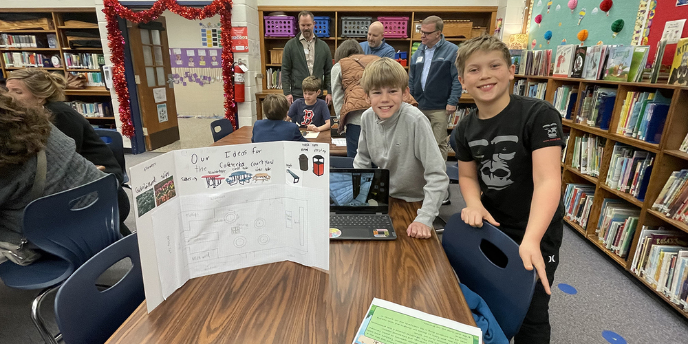 Students and adults gather in a school library around tables with laptops and a trifold project board, surrounded by bookshelves, posters, and classroom decorations.
