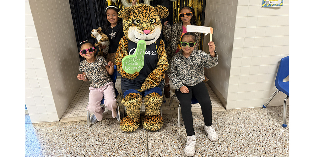 A spotted wildcat mascot wearing a dark shirt sits on a chair in a school hallway, surrounded by four children wearing sunglasses and leopard-print clothing. One child holds a green foam finger with “LCPS” on it, and another holds a small sign. Black-and-gold streamers hang in the background, and blue chairs line the tiled hallway.