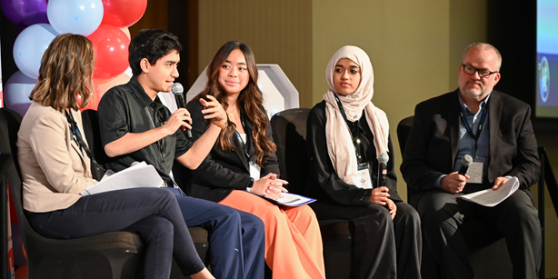 Five panelists seated on stage during a discussion, holding microphones and notes, with colorful balloons in the background.