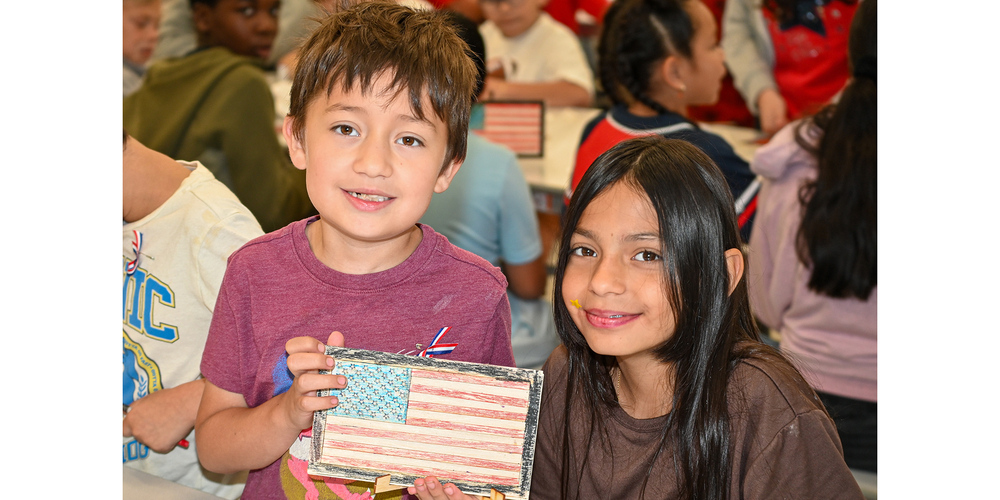 Two children sit at a classroom table and hold up a small American flag craft made from wood or cardstock with red, white, and blue coloring. Other students and classroom materials are visible in the background, slightly out of focus.