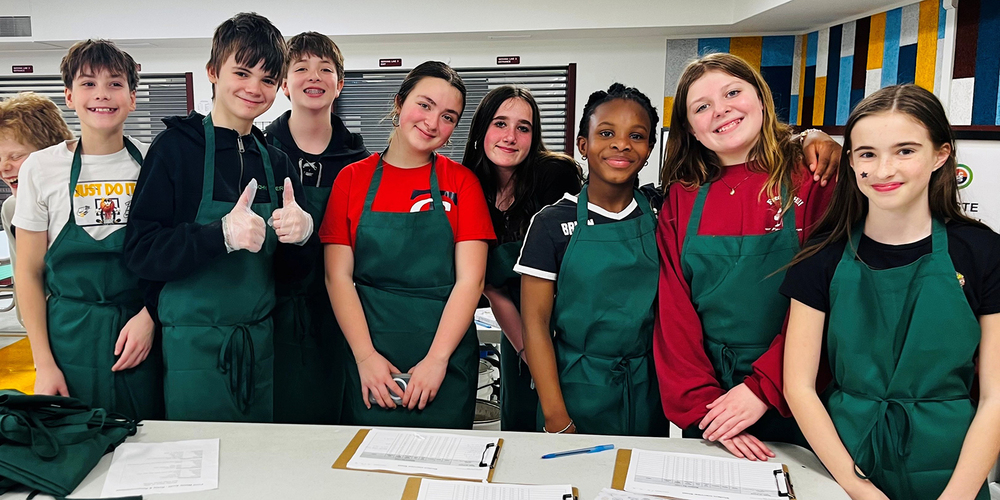 A group of students stands behind a classroom table wearing green aprons, posing together during a hands-on activity. One student in the group gives a thumbs-up gesture while others stand closely side by side. Clipboards with papers and a pen rest on the table in front of them, and a colorful panel wall and classroom equipment are visible in the background.
