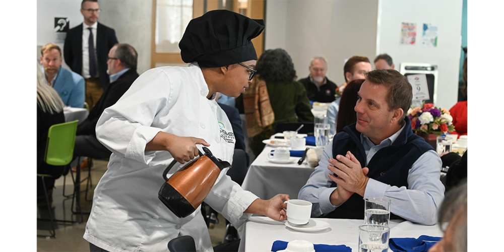 A server in a white chef coat and black hat pours coffee into a cup at a dining table with seated guests in a busy restaurant.