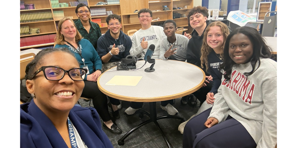 A group of adults and teenagers seated and standing around a round table in a library or media center. Audio recording equipment, including a microphone and headphones, sits on the table. Bookshelves filled with books line the background, and several people smile toward the camera while posing for a group photo.