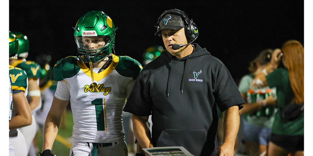 Football player wearing a green helmet and white jersey number 1 stands next to a coach in black hoodie labeled Coach Bishop during a night game.
