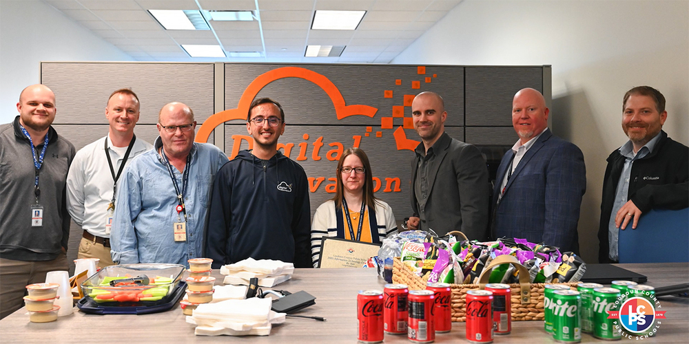 A group of nine people stand behind a conference table in an office, posing in front of a wall sign with a cloud logo and the words “Digital Innovation.” The table holds catered food, snacks, and soft drink cans, and one person near the center holds a certificate.