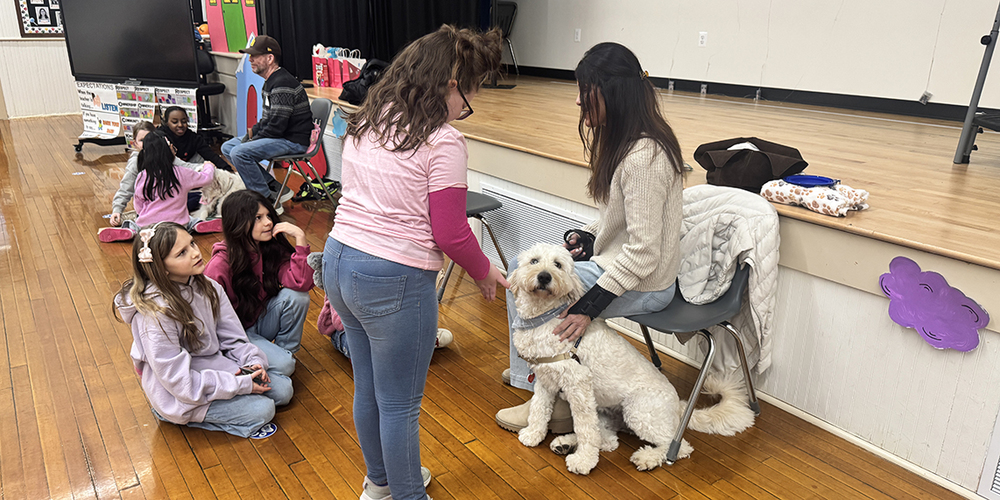 Children sitting on a wooden floor watching a woman seated on a chair petting a white dog near a stage in a gym.