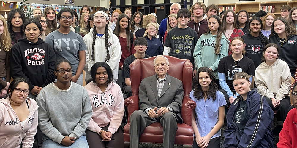 A Holocaust survivor sits in a red armchair surrounded by a large group of students posing for a group photo inside a school library.