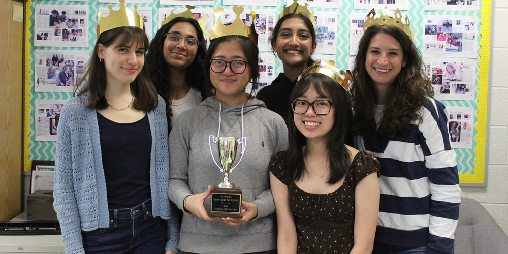 Group of six young people wearing gold paper crowns posing indoors with a trophy held by the person in the center.