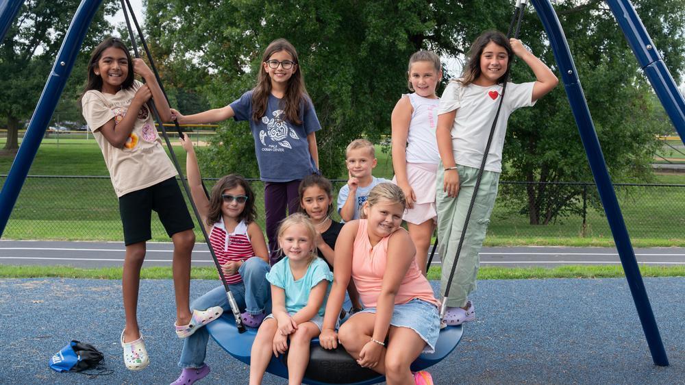LCPS children pose together on a large playground swing, smiling and gathered around the seat at a playground.
