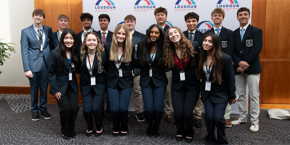 A group of approximately sixteen students stands and kneels in two rows at an indoor event, wearing coordinated business attire with name badges and school-style blazers. The group poses in front of a step-and-repeat backdrop displaying the word “Loudoun” with a red and blue logo, in a carpeted conference space with wood-paneled walls.