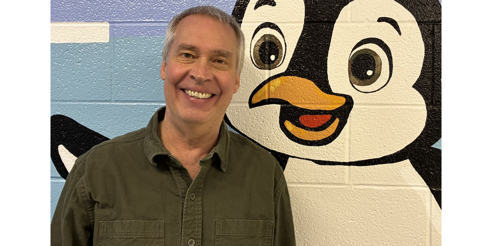 Alan Skubal in a green shirt standing in front of a cartoon penguin mural on a painted brick wall.