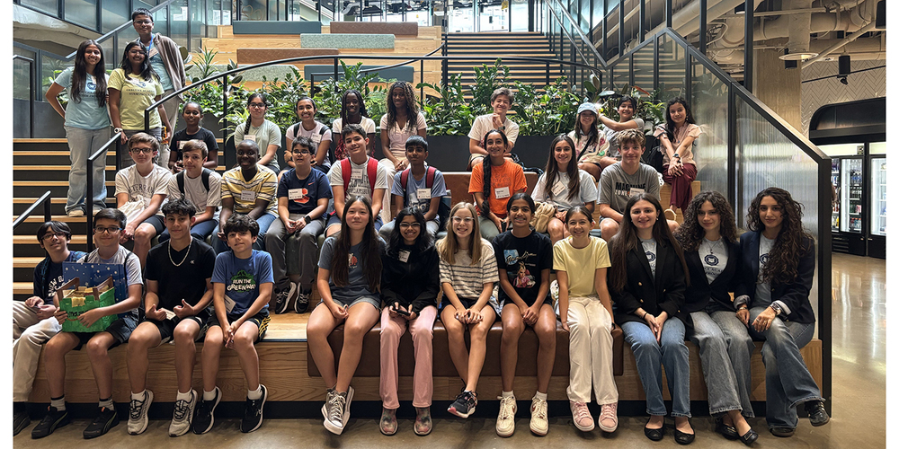 Group of students posing on tiered seating in a modern indoor space with staircases, glass walls, and large planters.