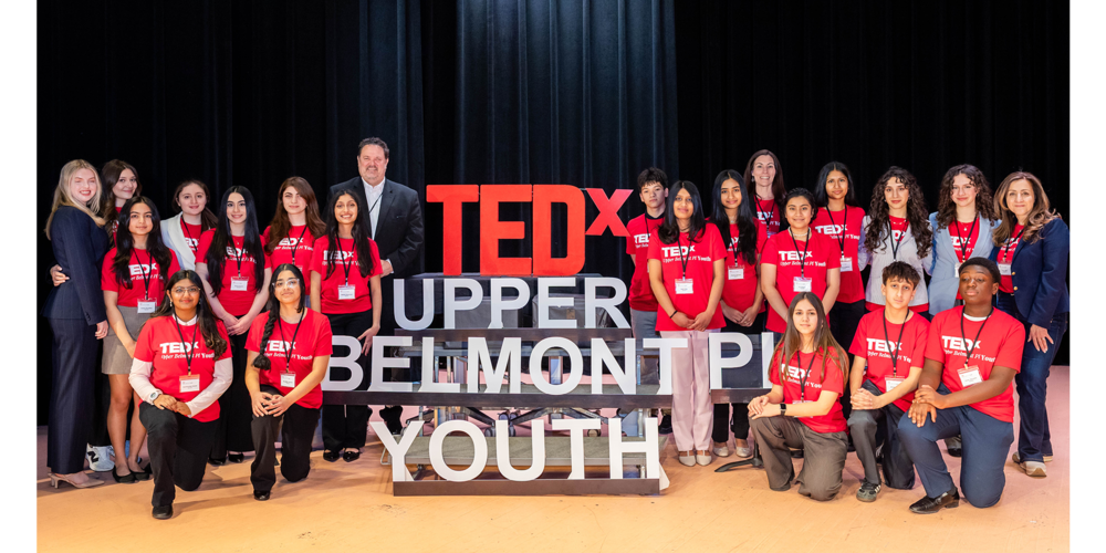 Group photo of diverse youth and adults wearing red TEDx Upper Belmont Place Youth shirts on stage with TEDx sign.