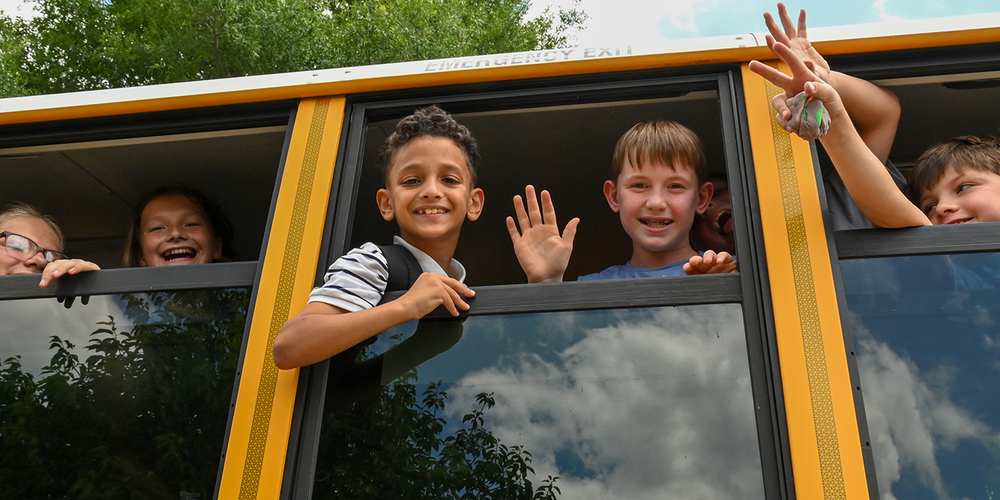 Several children sit by open windows on a yellow school bus, with arms raised and hands waving. The bus windows reflect clouds and trees, and green foliage is visible outside.