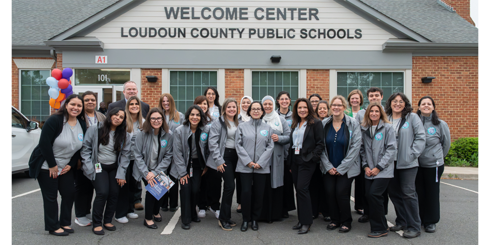 A large group of people wearing matching gray jackets stand together outside a brick building labeled “Welcome Center – Loudoun County Public Schools.” Red, white, and blue balloons are visible near the entrance as the group poses in front of the building during daylight.