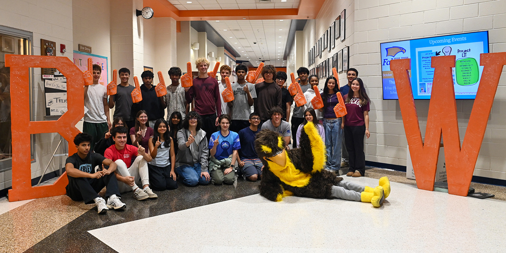 A large group of students poses in a school hallway between oversized orange letters “B” and “W.” Several students hold orange foam fingers and make a number-one gesture. A black-and-yellow bird mascot lies in front of the group on the floor. A digital display board and school posters are visible on the wall in the background.