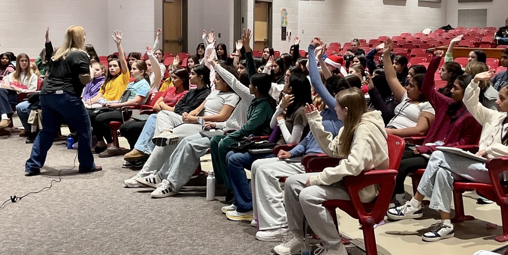 A classroom setting with students seated in red chairs, many raising their hands while a teacher stands at the front engaging them.