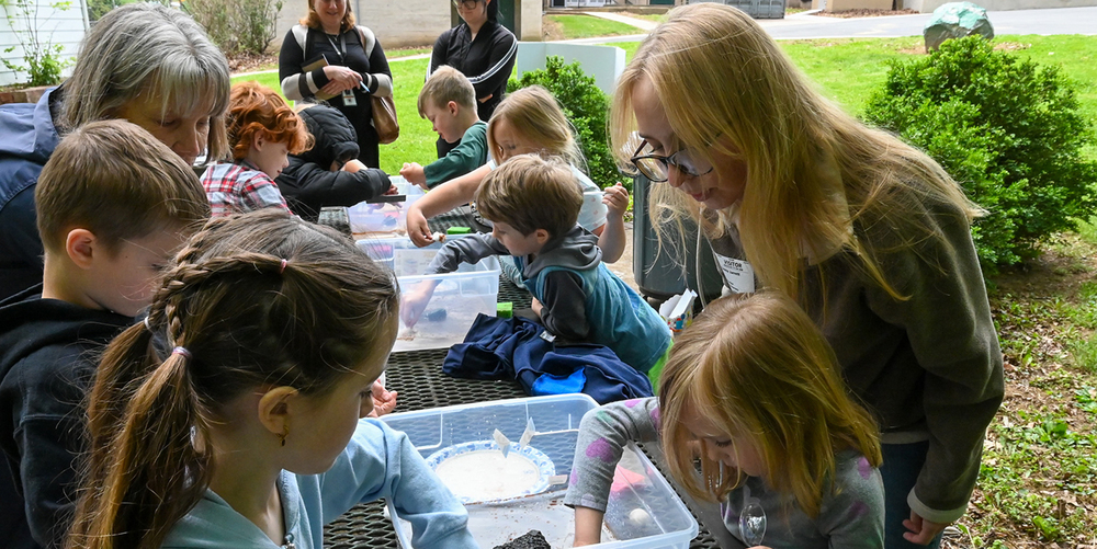 A group of children gathered around an outdoor picnic table, reaching into clear plastic bins filled with soil, rocks, and small materials for a hands-on activity. Several adults stand or lean nearby, observing and assisting. The scene takes place on a grassy area with shrubs and buildings in the background.