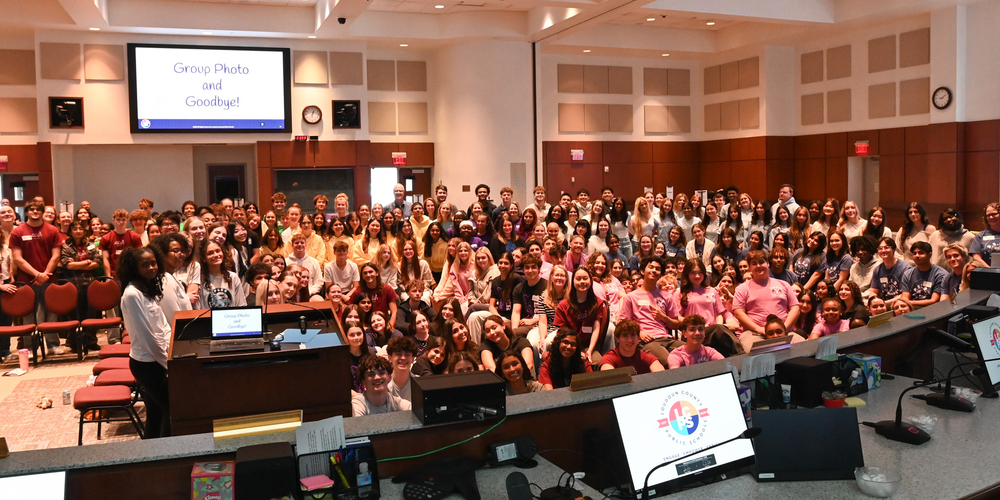 Large group gathered in the LCPS Board Room behind a desk with computers and microphones, with a screen reading “Group Photo and Goodbye!” displayed at the front.