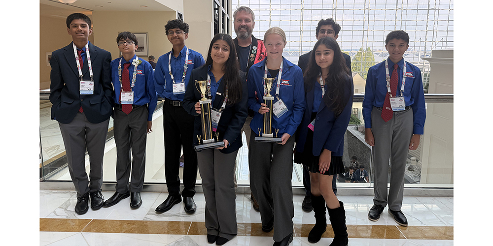 Group of students in matching academic attire posing indoors with two trophies in front of a large window and glass railing.