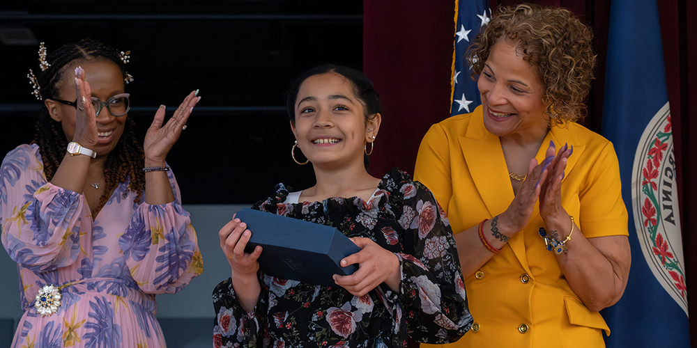 Two women and a female student stand on stage, one holding a box, while the others applaud, with flags in the background.