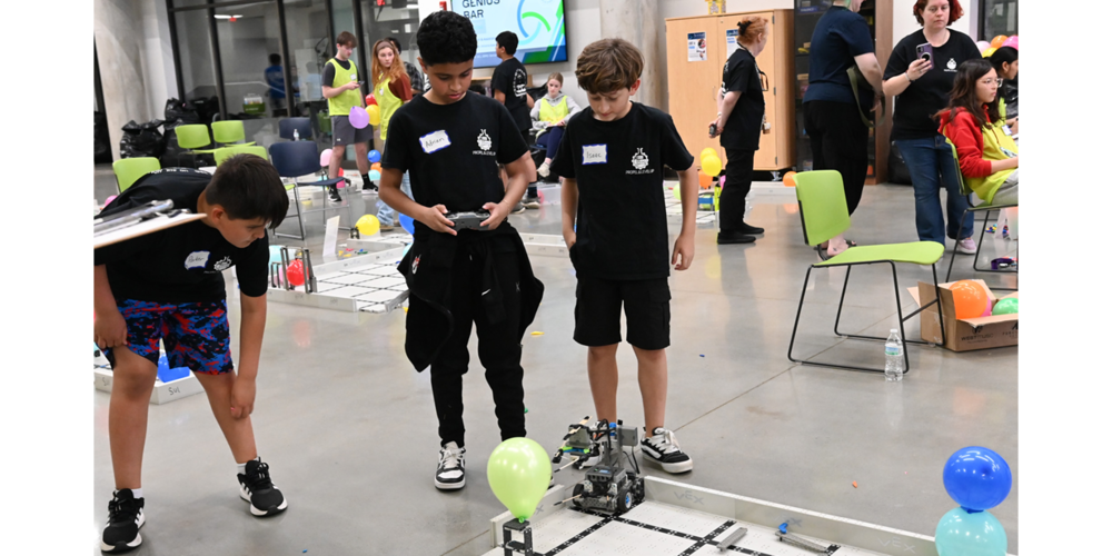Photo of students participating in a hands-on robotics activity inside a large indoor space. In the foreground, three students in matching black T‑shirts watch a small robot move along a taped grid on the floor, with balloons marking parts of the course. Other participants and seating are visible in the background.