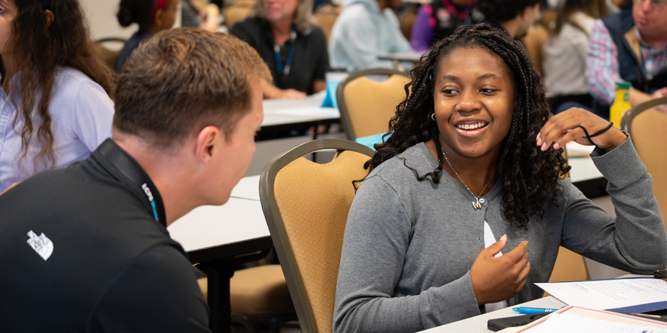 Two people seated at a conference-style table engage in conversation during a workshop or training session. One participant in a gray sweater gestures while speaking, with papers, a pen, and a smartphone on the table. Other attendees sit at tables in the background of the meeting room.