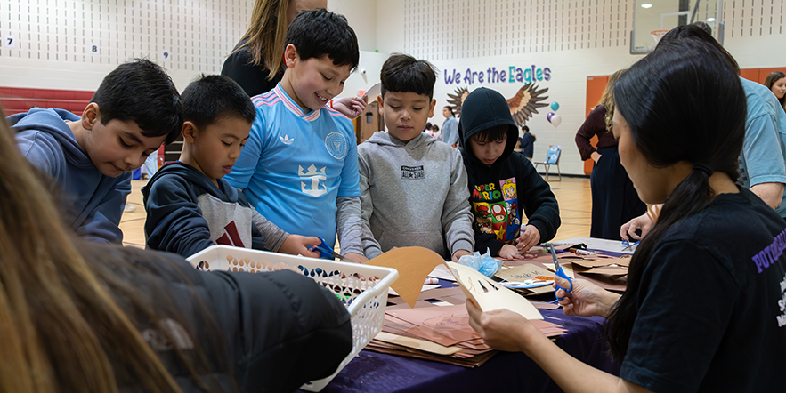 Children gathered around a table in a gymnasium, engaging in a group activity with paper bags and markers.