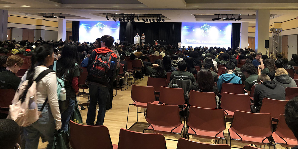 Large audience seated in a conference hall watching a speaker on stage with two large projection screens displaying presentation slides.
