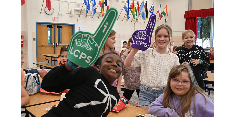 Several elementary-age students gather around cafeteria-style tables in a school multipurpose room decorated with international flags. Two students hold oversized foam “number one” fingers, one green and one purple, both with “LCPS” printed on them. Other students stand and sit nearby, with a backpack, snack bag, and lunch tables visible in the foreground. Double doors, windows with red curtains, and a wall clock appear in the background.