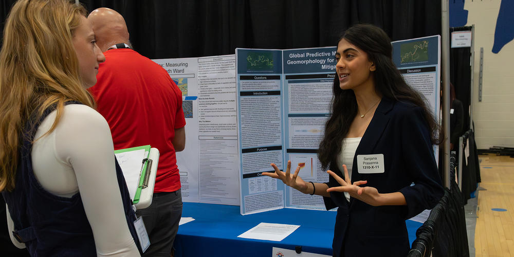 A female student standing beside a research poster presentation gestures while speaking to another attendee holding a clipboard. The poster display board behind her shows charts and text, and additional conference attendees and displays are visible in a gymnasium-style venue.