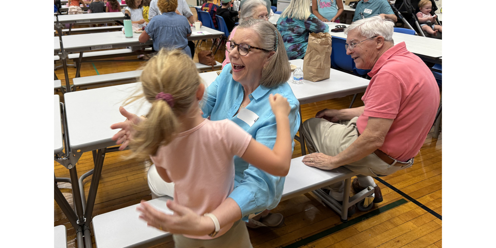 A child with a ponytail approaches an older adult seated on a cafeteria bench with outstretched arms, while another older adult sits nearby. The scene takes place in a busy community hall or school cafeteria with long tables, benches, and families gathered in the background.