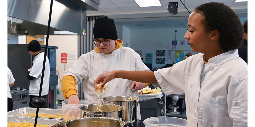 Two chefs in white uniforms preparing food together in a professional kitchen with large pots and cooking utensils.