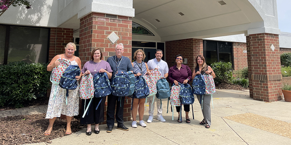 Seven adults stand in front of a brick office building entrance, each holding backpacks and colorful patterned tote bags. The group is lined up on a sidewalk beneath a covered entryway with brick columns and large windows. Shrubs and landscaping border the building, and the backpacks appear to be part of a donation or community-support effort.