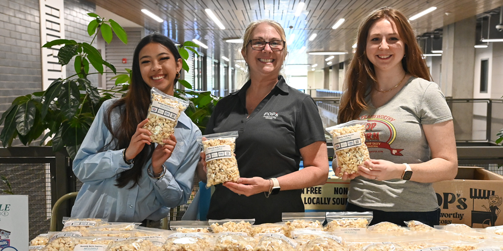 Three people stand behind a table covered with packaged popcorn in a modern indoor space with plants and overhead lighting, each holding a labeled bag of popcorn toward the camera.