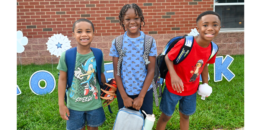 Three school-aged children wearing backpacks stand on a grassy area in front of a brick school building. Decorative blue letters spelling part of “BACK TO SCHOOL” and paper starburst decorations are displayed behind them. One child holds a lunch bag and water bottle, while another carries a white pom-pom-like item. The children face the camera outdoors on a bright day.