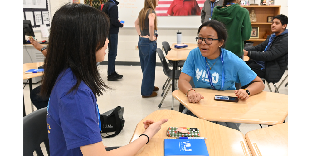 A teacher and student engaged in conversation at a classroom table, one wearing a blue shirt and the other with long dark hair in a purple shirt.