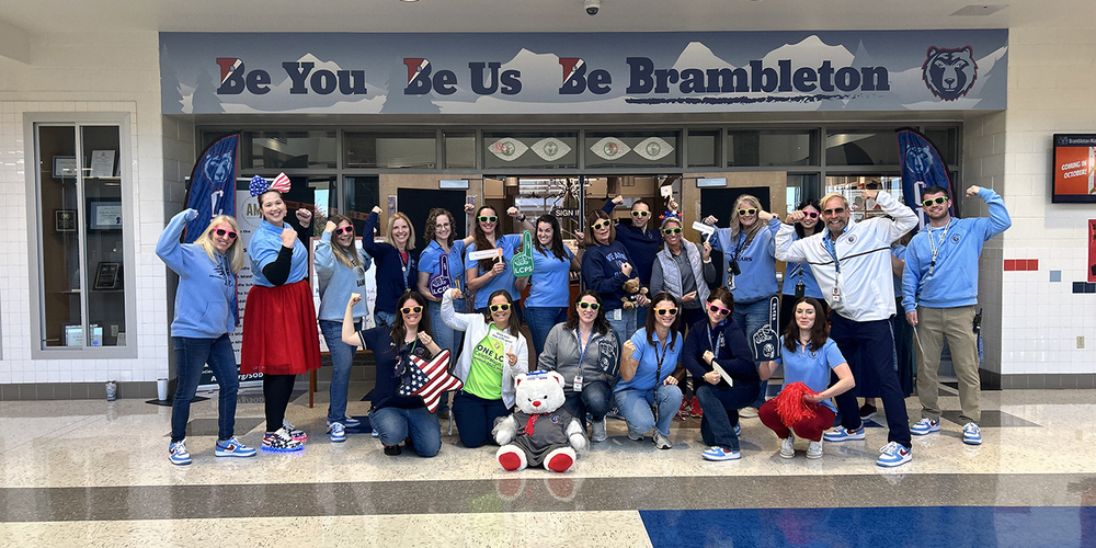 Group of adults posing energetically in front of a school entrance with a 'Be You Be Us Be Brambleton' banner overhead.