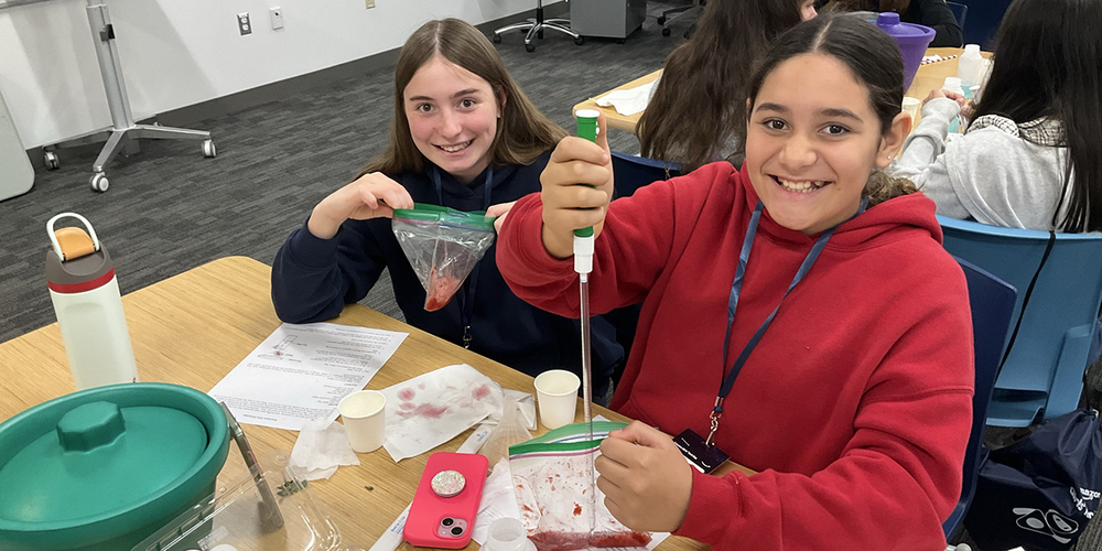 Two people sit at a classroom table doing a hands-on science activity. One person uses a pipette to transfer a red liquid into a clear plastic bag while the other holds a similar bag; cups, papers, a water bottle, and lab supplies are spread across the table, with other participants working in the background.