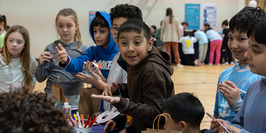 Children gathered around a table in a gymnasium working on arts and crafts with paper, markers, and glue, while more children participate in activities in the background.
