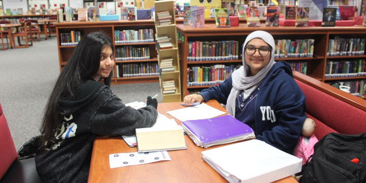 Two students study together at a library table surrounded by bookshelves filled with books.