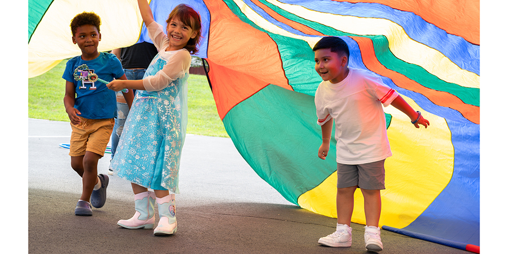 Three children play beneath a large multicolored parachute outdoors. One child in a blue shirt and shorts stands at left, another in a light blue dress and boots holds the parachute overhead in the center, and a third child in a white shirt and gray shorts runs underneath on the right. The bright orange, blue, green, and yellow fabric fills much of the scene, creating a colorful canopy above a paved play area.