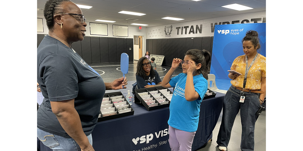 A student tries on a pair of eyeglasses at a VSP Vision Eyes of Hope vision-screening event inside a school gymnasium. An adult standing nearby holds a hand mirror while the student adjusts the glasses and looks at the reflection. Tables covered with VSP Vision tablecloths display trays of eyeglass frames, and additional staff members stand behind the tables. Wrestling mats and school signage are visible in the background.