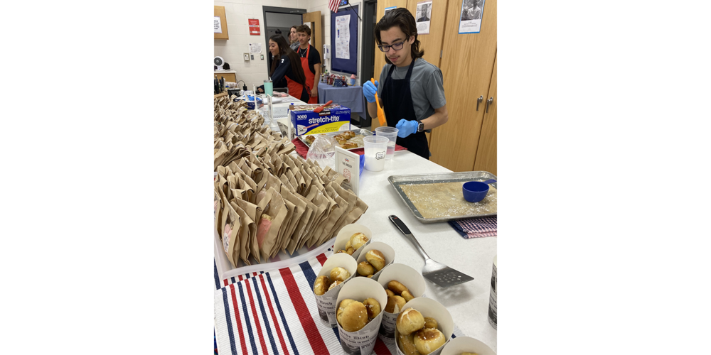 Students wearing aprons and gloves prepare food in a school kitchen with rows of packed brown paper bags on the counter.